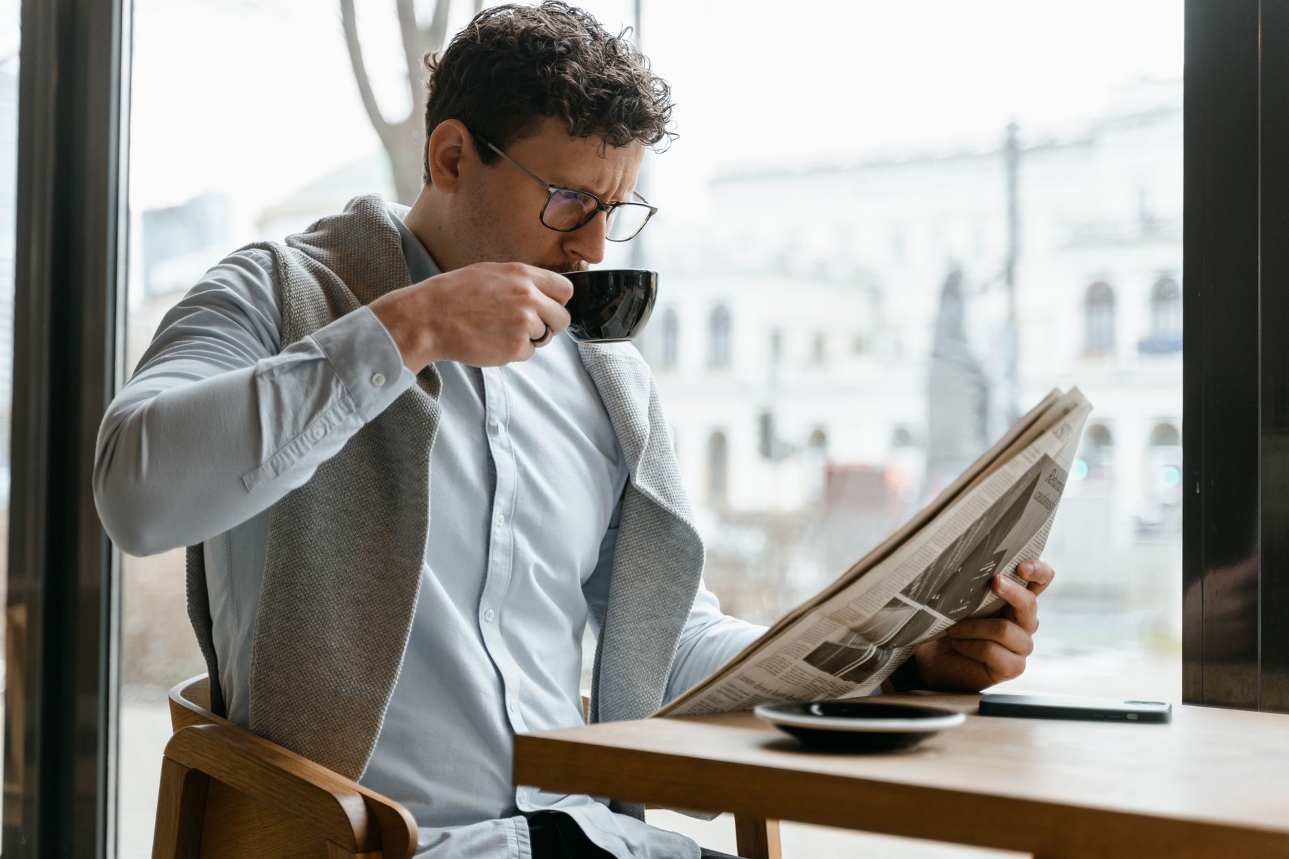 Adult man wearing eyeglasses sipping coffee and reading a newspaper in a cozy cafe setting. — Gama Casino Erfahrungen: Spiele